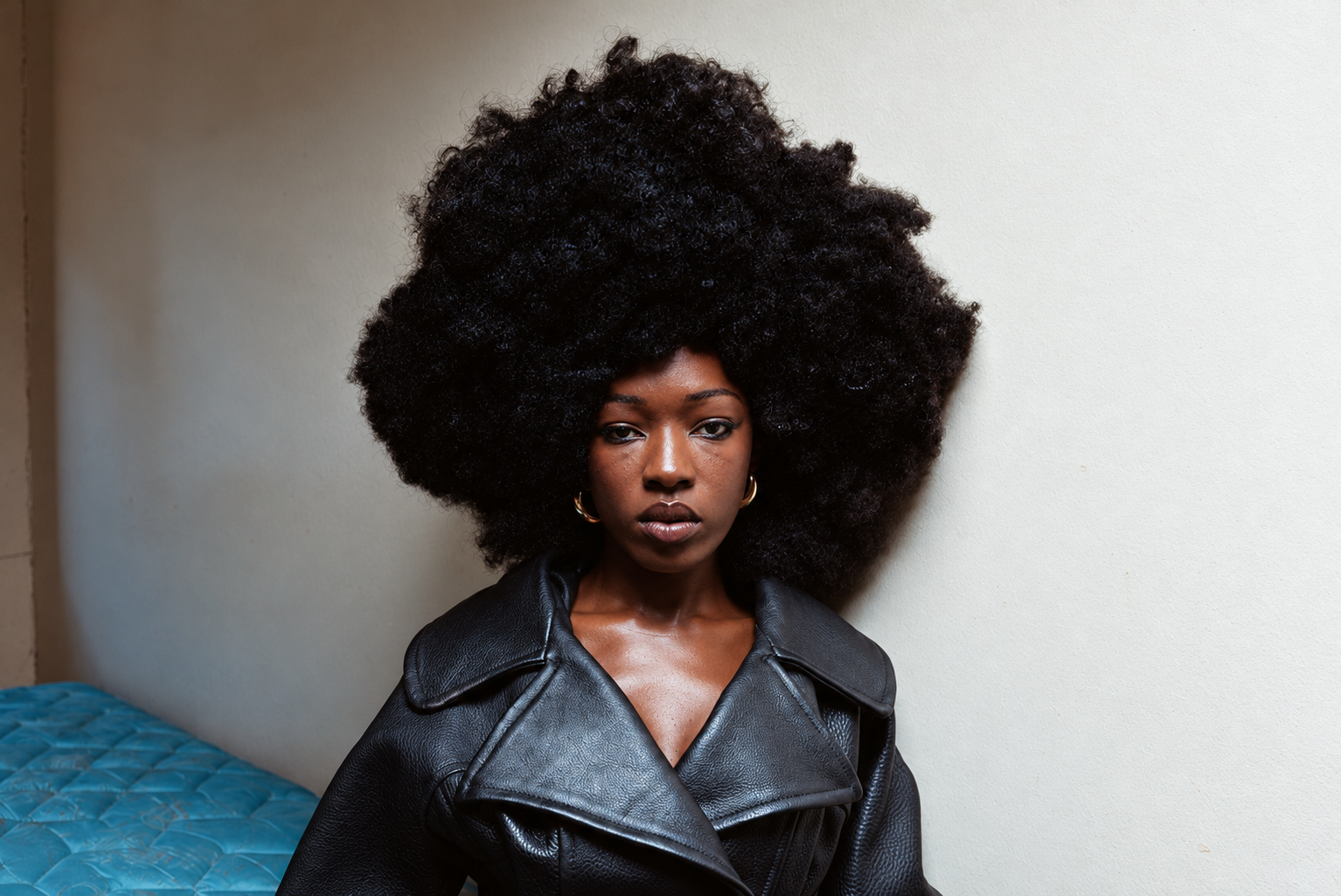 Elsa Majimbo en manteau noir, assise au bord d’un lit, avec une coiffure afro