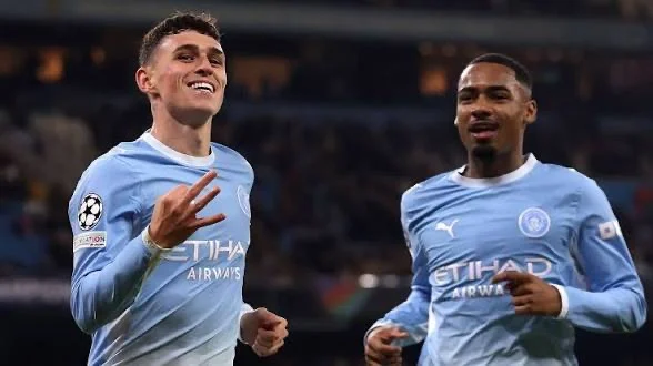 Deux joueurs de Manchester City en tenue bleu ciel, dont un montre trois doigts.