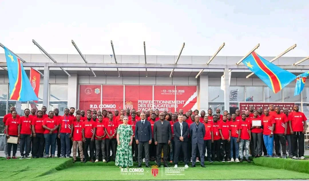 Academy coaches in red T-shirts pose with officials in front of the Stade Tata Raphaël building, between DR Congo flags.