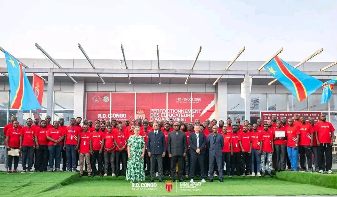 Les éducateurs d’académies, en t-shirts rouges, posent avec les officiels devant le bâtiment du stade Tata Raphaël, entre des drapeaux de la RDC.